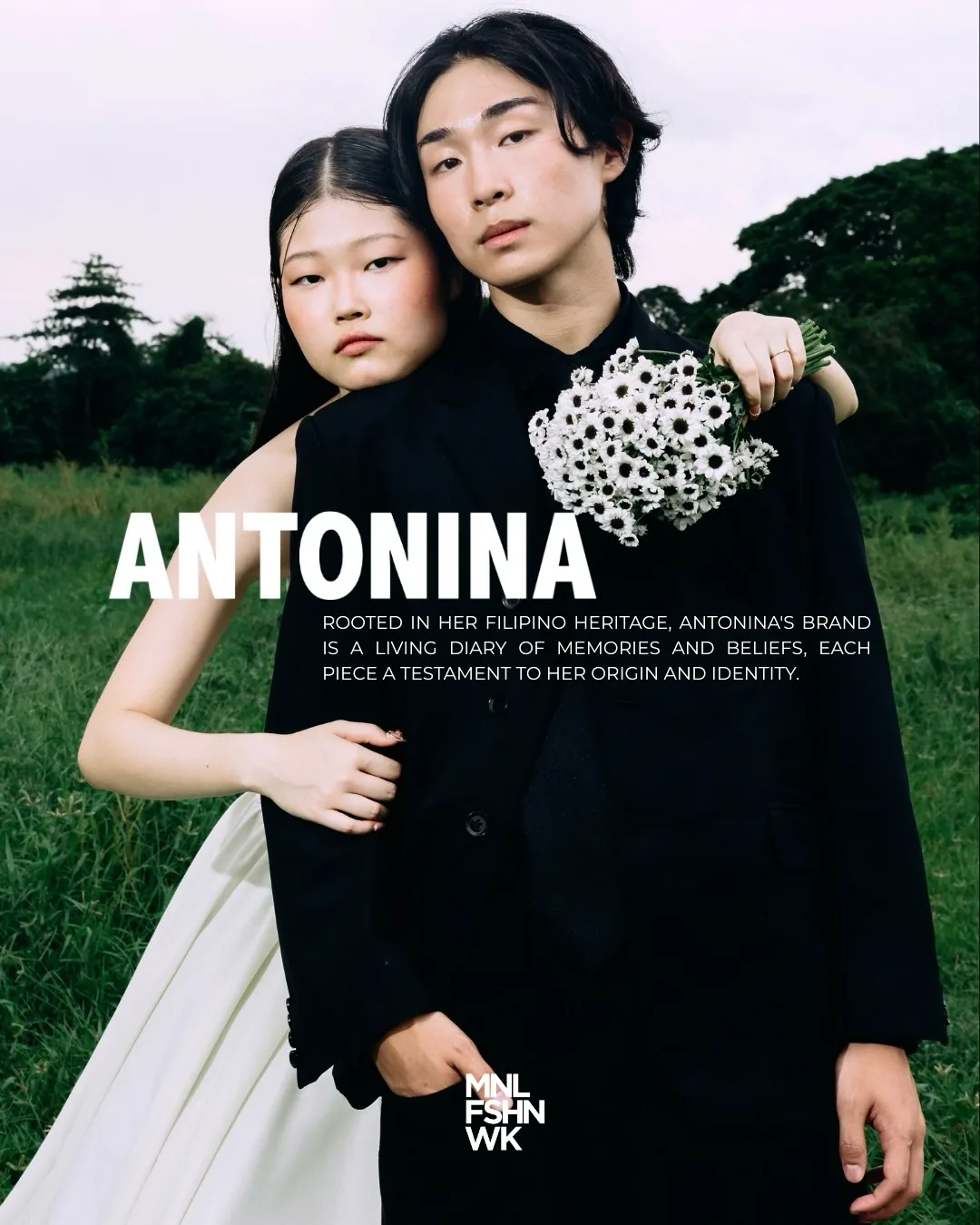 Two Antonina models embracing in a field, the bride's ivory train catching the wind beside a black tailored suit with a white flower boutonniere